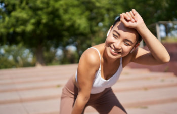 A woman wearing a white tank top and brown pants is standing outdoors, smiling and leaning forward with one hand on her knee and the other on her forehead. Trees and steps are visible in the background.